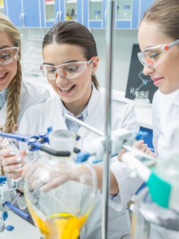 Smiling female scientists in protective eyewear making experiment in chemical laboratory